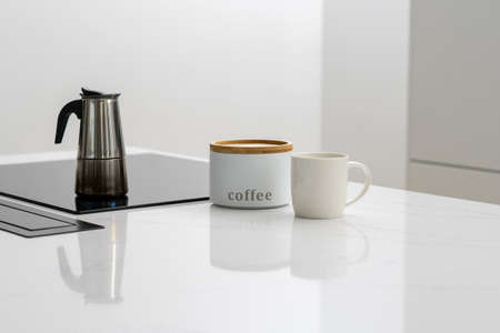 Brewing coffee for breakfast in steel pot on built in induction ceramic cooktop, white labeled jar containing coffee with mug next to it in kitchen with minimalistic designの写真素材