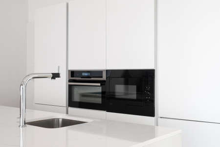 White glossy countertop with shiny stylish metal faucet and sink in clean modern minimalist kitchen with black oven and microwave built in big cupboardの写真素材