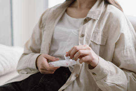 Concept of daily and weekly pill intake. Cropped view of young woman holding organizer with medicament from mental disorder disease, depression and seasonal allergy. Ill female patient stay at homeの写真素材