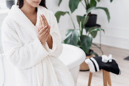 Cropped shot of woman model in white bathrobe touching soft skin on her hands and applying cream while sitting on bathtub in spacious light bathroom with green plant on background. Body care conceptの写真素材