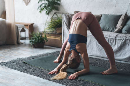 Hobby and lifestyle concept. Young adult woman in sportswear lean down, warming up, stretching legs, touching exercise mat with her hands and making daily sport training at homeの写真素材