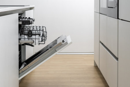 Kitchen with white furniture and built in electrical devices, open dishwasher with basket and clean tableware inside, dishes and crockery laying after being washed by machineの写真素材