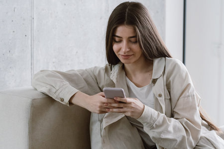 Happy young woman in casual clothes using online app at cellphone, sitting on comfort couch in...