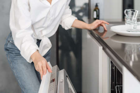 Housewife in casual clothes opening door of modern dishwashing machine after finished cleaning process while doing daily housework at homeの写真素材