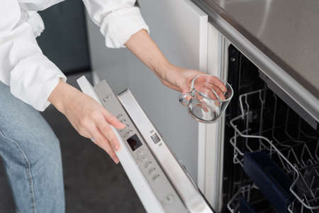 Cropped shot of female washing dishware in modern dishwasher while cleaning in kitchen at home, putting glass inside. Housework and household chores conceptの写真素材