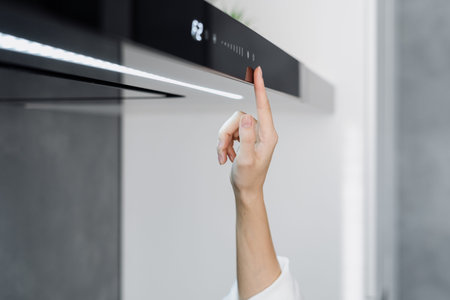 Cropped shot of woman using cooker hood in minimalist modern kitchen with modern integrated appliances, female pressing button on cooking extractor while preparing food at homeの写真素材