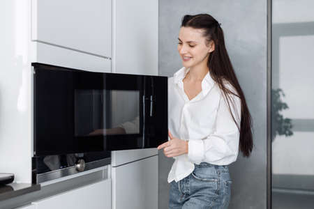 Happy smiling woman in casual outfit opening black microwave door and going to put food in to heat it up while standing in stylish kitchen with white furniture at homeの写真素材