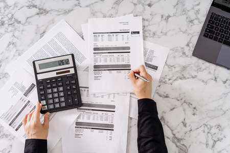 Cropped shot of woman calculating important monthly expenses, estimating total utility payments, using calculator and laptop, table with marble surface, writing with pen on paperの写真素材