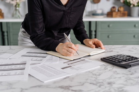 Woman in black shirt writing total cost in note book after calculating expenses and bills using calculator, kitchen environment with vintage green furniture in blurred background. Cropped shotの写真素材