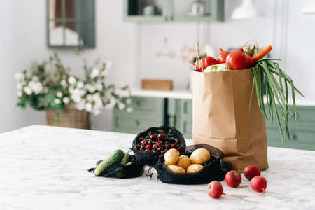 Various fresh vegetables in paper grocery and black mesh bags on kitchen island with marble top, healthy food full of nutrients, vintage furniture with flowers, blurred backgroundの写真素材