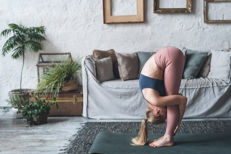 Full length of sporty female in active wear standing in Uttanasana pose while practicing yoga at...