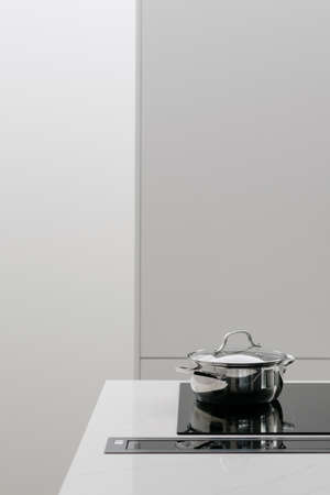 Metal pot with food being cooked on glass electric stove with retracted ventilation strip built in white kitchen counter, minimalistic style and simple furniture. Vertical shotの写真素材