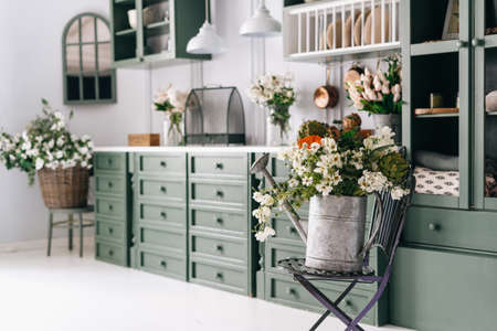 Kitchen with green and white theme, old metal watering pot full of greenery on folding chair, flowers all over counter in blurred background, vintage monochrome furnitureの写真素材