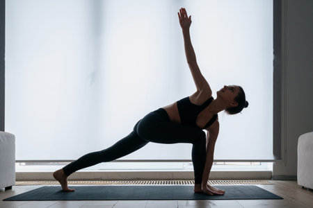Silhouette of young woman exercising at home, doing revolved crescent lunge with arms extended yoga...