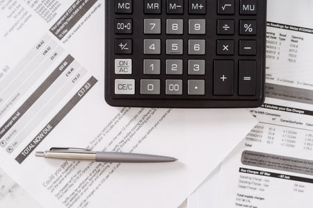 Top view of black calculator with pen laying on various invoices and monthly utility bills on table with paper scattered aroundの写真素材