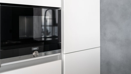 Spacious white cupboard with built-in glossy black microwave oven, control knob to adjust power and mode, display to show time left, modern kitchen appliancesの写真素材