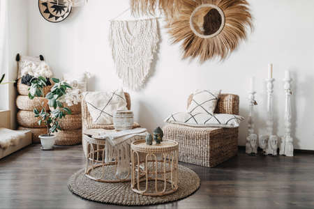 Home decor concept. Comfortable wicker furniture, rattan armchair with cushions, bamboo coffee table and macrame on white wall in cozy living room with ethnic interior designの写真素材