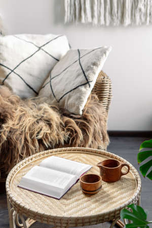 Concept of recreation, relaxation. Weekend at cozy house. Wicker coffee table with open book and wooden cup with beverage standing near rattan armchair. Living room interior at boho chic styleの写真素材