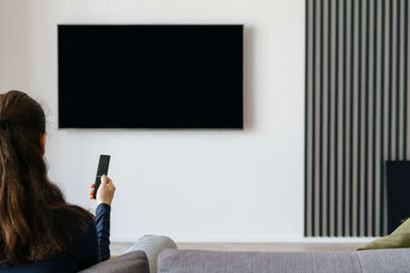 Rear view of woman resting on couch holding remote in front of mock up TV hanging on white wall in cozy apartment. Living room of modern houseの写真素材