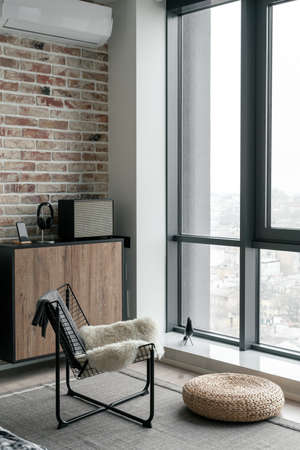 Vertical shot of relax area with metal chair and straw ottoman opposite bright panoramic window, wooden commode with modern sound bluetooth column, headphones and smartphoneの写真素材