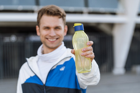 Selective focus on bottle with drink water. Happy and smiling sportsman holding holding protein shake with vitamins in hand, standing after sport training outdoorsの写真素材