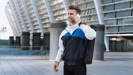 Side view of confident man with yoga mat behind his back talking at modern smartphone before sport training in city. Sportsman prepare to daily workout, standing outdoorの写真素材