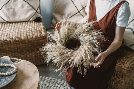 Cropped woman holding handmade braided wreath of dried plants in boho chic style, sitting on wicker armchair in florist studio. Female working at flower shop with eco materialsの写真素材