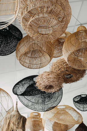 Low angle view of many handmade and wicker lampshade with natural materials hanging in showroom. Vertical view of lighting equipment in boho chic styleの写真素材