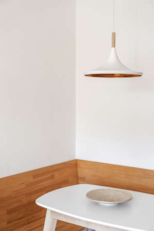 Vertical view of white and modern ceiling electric lamp hanging over kitchen table with empty wooden bowl. Morning in modern room at contemporary apartment with minimalist interior designの写真素材
