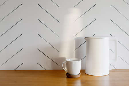 White and modern electric teapot and cup for coffee or herbal tea on wooden table against white copy space wall. Concept of cozy kitchen and morning beverageの写真素材