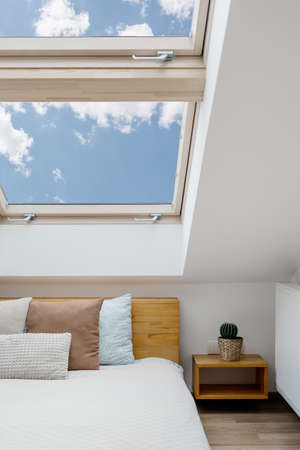 Concept of morning light in house. Vertical view of clean skylight in white bedroom interior. Modern apartment with attic window in small room with home decor, green house plants and cushions on bedの写真素材