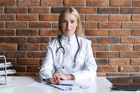 Healthcare concept. Confident family doctor looking at camera, sitting behind table in private clinic office. Qualified GP wearing in white medical uniform with stethoscope at workplace in hospitalの写真素材