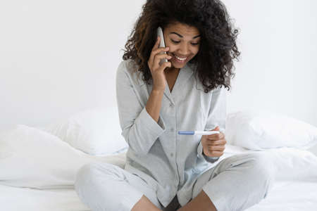 Maternity concept. Happy african american woman holding pregnancy test, talking on smartphone, sharing good news with husband or her boyfriend, sitting on bed in sleepwear and smiling wideの写真素材