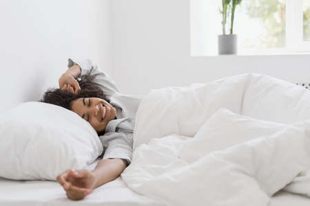 Healthy sleep, recreation and relaxation concept. Happy young afro american woman waking up in white bedroom, stretching arms, lying on bed in sleepwear. Millennial girl spend early morning at homeの写真素材