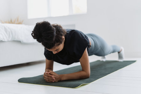 Focused african ethnicity woman in sweatpants and t-shirt standing in plank position on yoga mat,...