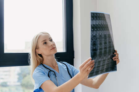 Qualified nurse or focused physician checking mri scan, looking at x ray picture, diagnostic disease of patient, standing in private clinic in medical uniform with stethoscopeの写真素材