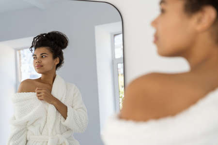 Calm and beautiful young afro american woman standing near mirror in bathroom wearing in bathrobe, showing naked shoulder, looking aside and smiling nice. Natural beauty, body and skin care conceptの写真素材