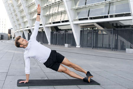 Sportsman training outdoors, raised hand up, standing in side plank pose working out on yoga mat. Sporty man in sportswear making abs or balance exercise, full lengthの写真素材