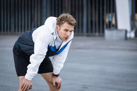 Side view of tired runner breathing fresh air after jogging on stadium, resting before daily hard training outdoors. Exhausted sportsman warming up in city, standing on urban copy space backgroundの写真素材