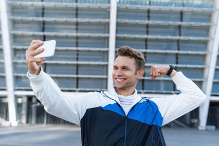 Concept of online workout with coach. Side view of happy athlete take selfie photo on smartphone after training outdoors. Man raises arm, show biceps and use camera, recording video after morning runの写真素材