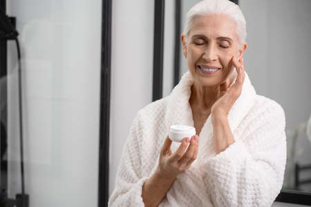 Portrait of aged woman with jar of cream in hands. Complexion face clearing process. Usage of anti-aging cosmetics. Skincare treatment with serum conceptの写真素材