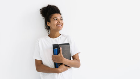 Young happy student girl looking aside, holding books and notebook in hands and standing against white background with blank space for education advertising. Learn, knowledge, study conceptの写真素材