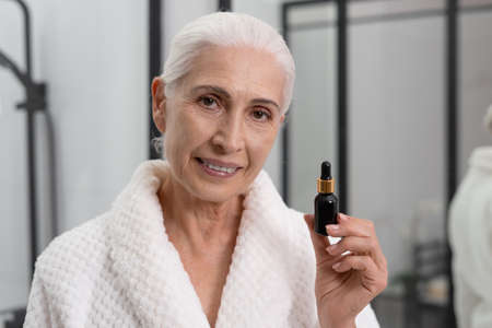 Portrait of elderly woman looking at camera and holding vial of serum. Skin care at home. Usage of anti-aging cosmetic procedures. Facial rejuvenationの写真素材