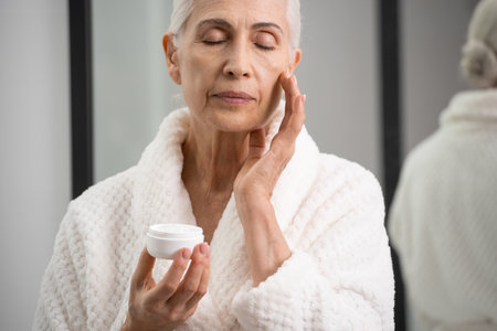 Portrait of elderly woman with bottle of cream in hands touching her face. Complexion clearing. Usage of anti-aging cosmetics. Skincare treatmentの素材 [FY310178538441]