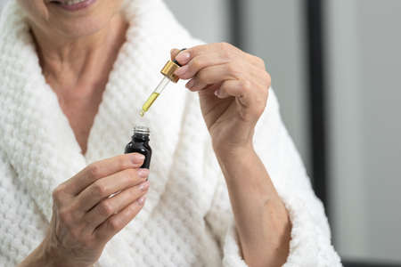 Closeup of elderly woman in bathrobe with dropper and bottle of serum in hands. Face skin care at home. Body rejuvenation procedures. Cosmetology and dermatology conceptの写真素材
