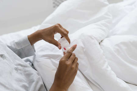 Cropped view of afro american woman holding box with pills in hands, sitting in bed, wearing in sleepwear. Sick female taking medicines at home. Healthcare, health problems conceptの写真素材