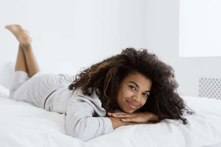 Portrait shot of joyful and charming afro american woman looking at camera, smiling nice, lying on bed in sleepwear. Candid shot of young female spend morning in bedroom, enjoying weekend or day offの写真素材