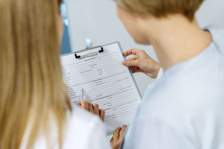 Back view of professional female dentist standing in stomatology clinic, showing health insurance to patient, fill form, making notes in document, discussing a treatment planの写真素材