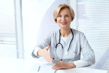 Smiling and confident family doctor sitting behind table in private clinic office, reaching hand for handshake, looking at camera. Concept of trust, support and patients health careの写真素材