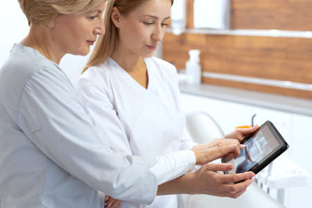 Side profile view of two professional dentist checking x ray picture on digital tablet, discussing patient treatment plan, stand in stomatology clinic. Concept of extraction and prosthetics of teethの写真素材
